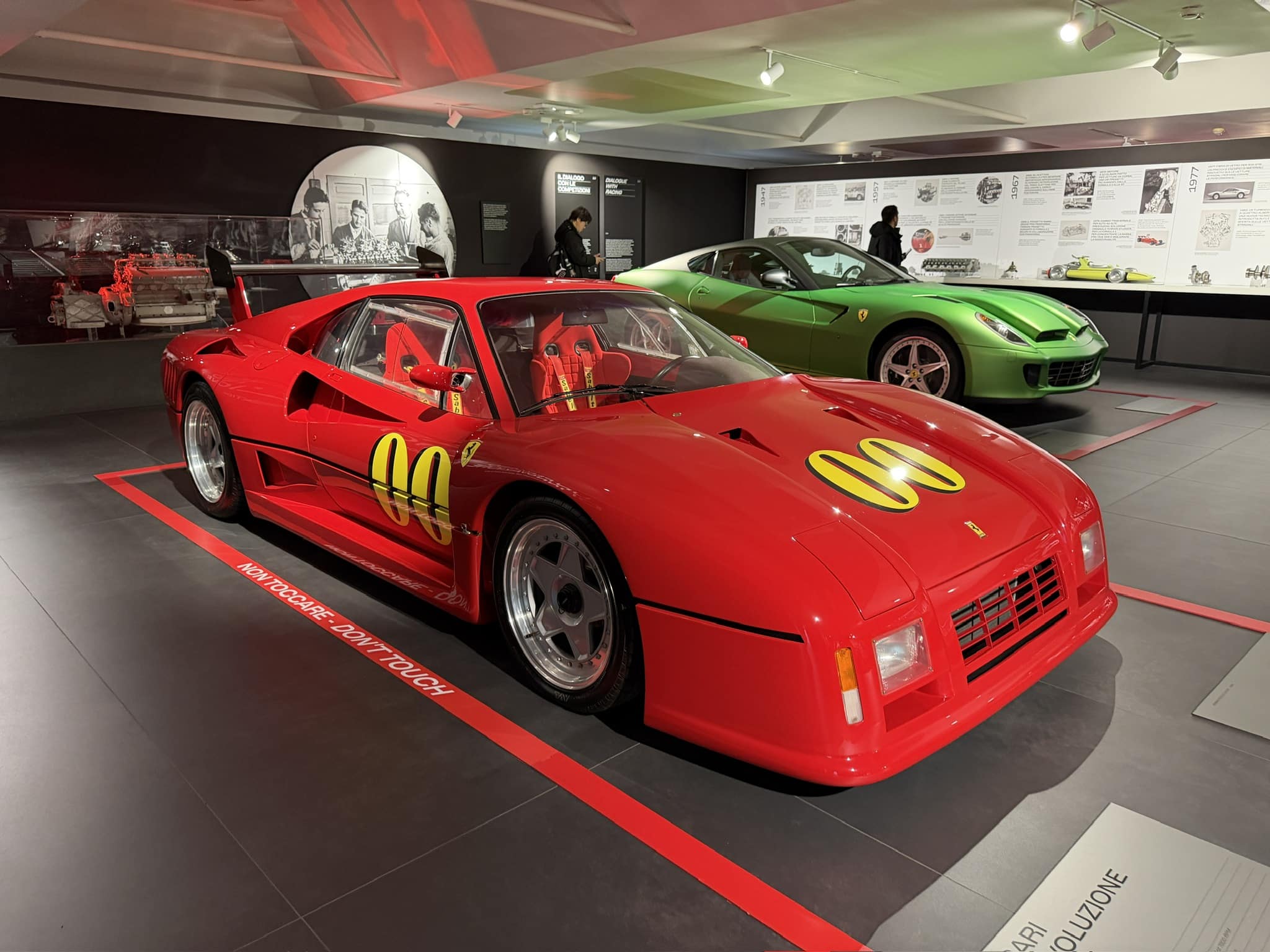 A sleek red Ferrari race car with number 90 sits displayed alongside a green sports car in a modern automotive museum