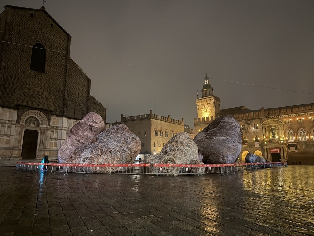 Large stone sculptures displayed in historic Italian piazza at night with illuminated medieval buildings and clock tower.